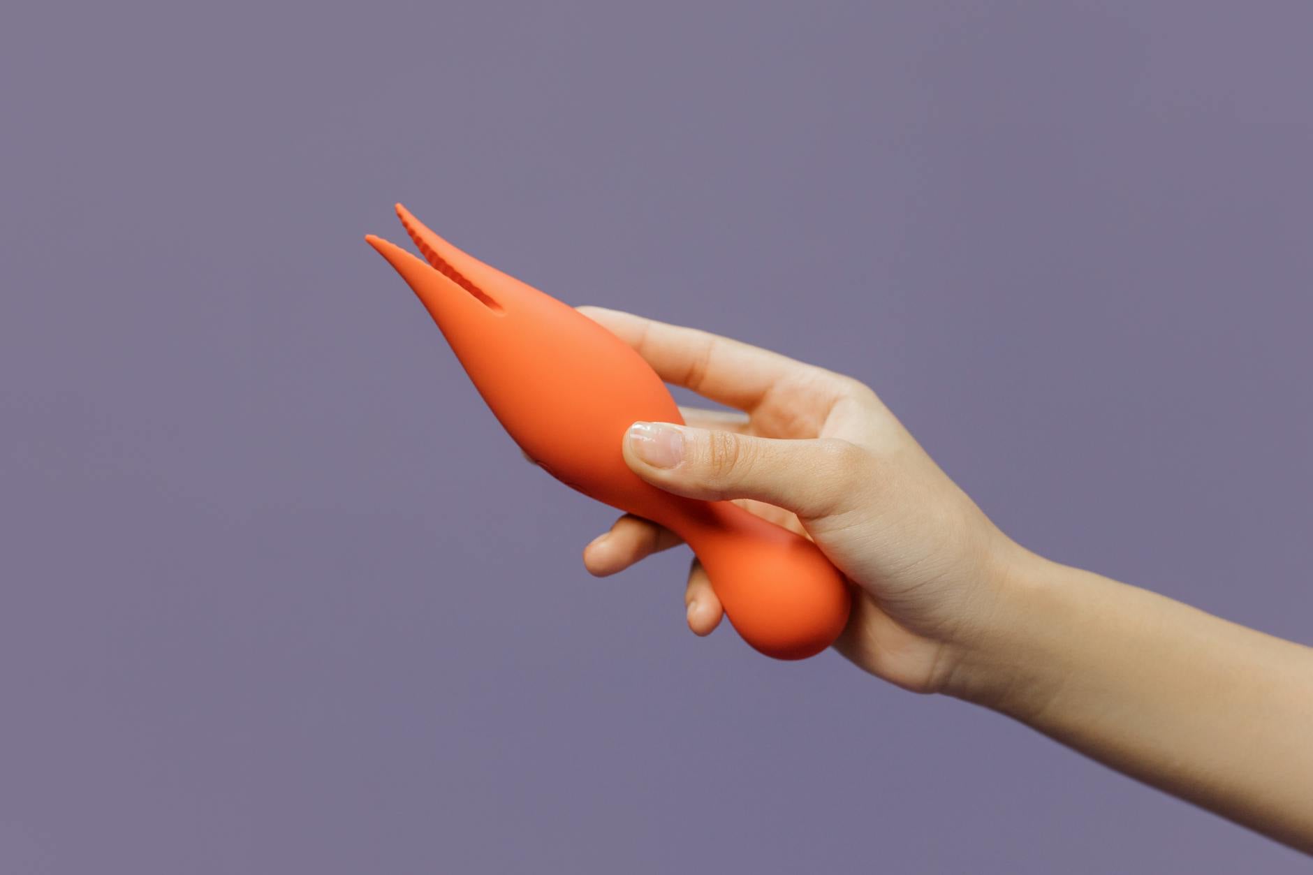 A hand holding a bright orange vibrator against a minimalistic purple backdrop, representing the return of pleasure and self-discovery.