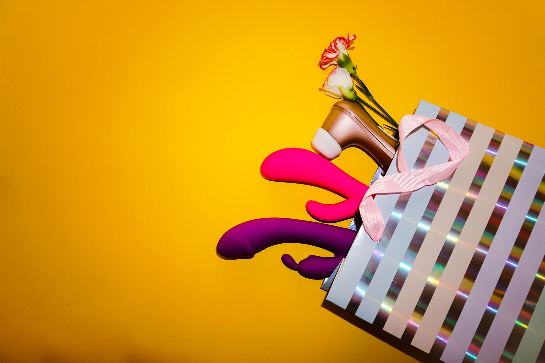 Colorful vibrators and clitoral toys displayed in a holographic gift bag against a bold yellow background